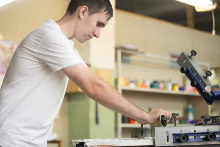 Young attractive worker man working using printmaking tools, silk screen printing on clothing fabric technique